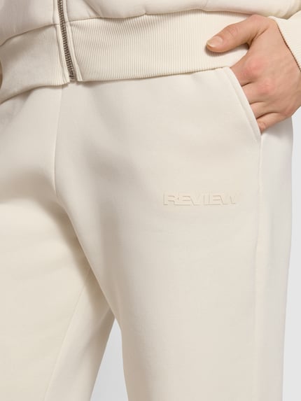 Vrouwenmodel draagt Basic Sweatpant met boordjes aan de zoom en zijzakken van review, katoenmix, los model, labelprint, geborduurd logo, detailopname.