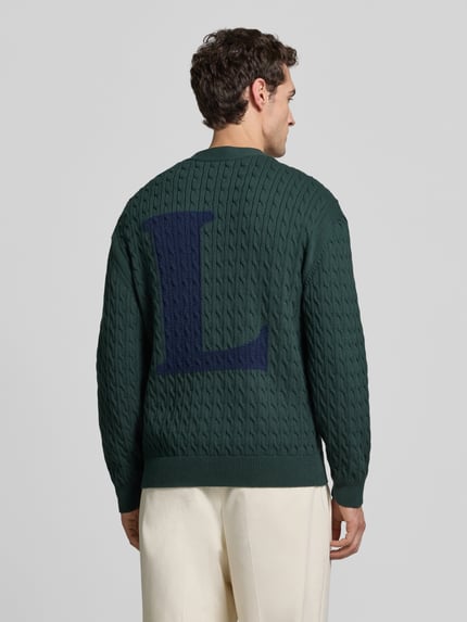 Männermodel trägt Cardigan mit Logo-Detail von Lacoste in dunkelgrüner Strickoptik mit Zopfmuster, gerippten Abschlüssen, Ganzkörperansicht Rückansicht.