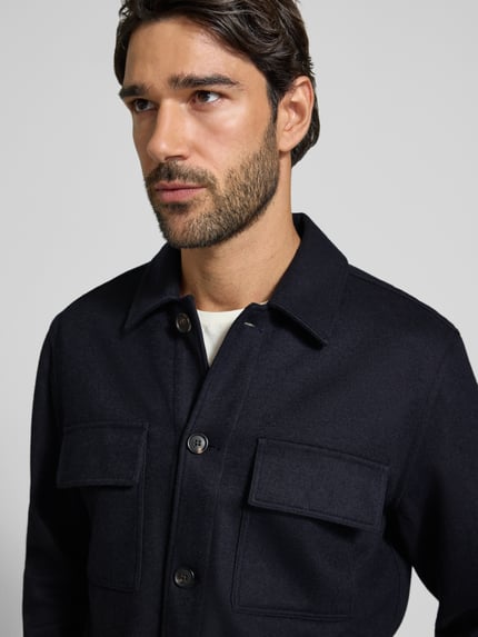 Męski model nosi Regular Fit Hemdjacke mit Kentkragen und Brusttaschen od Profuomo, materiał z domieszką wełny dziewiczej, ciemnogranatowy kolor, guziki i kieszenie na piersi, krój regular fit, widok na górną część ciała z przodu.