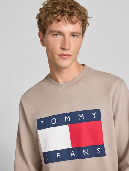 Mężczyzna nosi Regular Fit Sweatshirt aus reiner Baumwolle von Tommy Jeans, beżowa bluza z dużym logo na piersi, zbliżenie górnej części ciała.