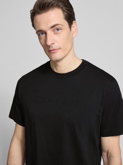 Männermodel trägt T-Shirt mit Rundhalsausschnitt von CK Calvin Klein, schwarze Farbe, Reine Baumwolle, Regular-Fit, Logo, Nahaufnahme Oberkörper. Rückenlänge bei Größe M: 67 cm.