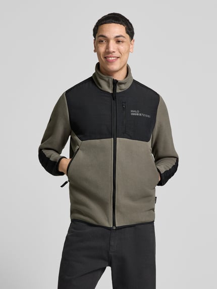 Męski model nosi Fleecejacke mit Stehkragen und Reißverschlusstaschen od HALO w kolorach khaki i czarnym, nadruk z logo, krój regular fit, stójka, zamek błyskawiczny, kieszenie zapinane na zamek błyskawiczny, długość tyłu w rozmiarze M: 71 cm, szerokość ramion w rozmiarze M: 45 cm, szerokość klatki piersiowej w rozmiarze M: 45 cm, długość rękawów w rozmiarze M: 69 cm, zdjęcie całej sylwetki z przodu