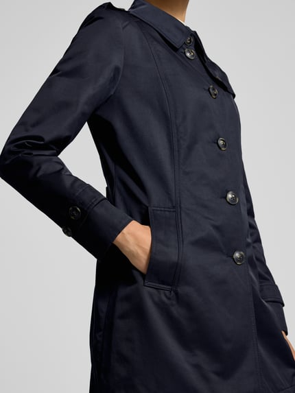 Frauenmodel trägt Trenchcoat mit Ärmelriegeln von Fuchs Schmitt in Dunkelblau, sichtbare Knopfleiste und Eingrifftaschen, Nahaufnahme Oberkörper.