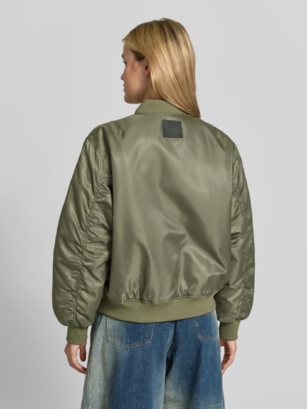 Frauenmodel trägt Bomberjacke mit Ärmeltasche Modell 'LEON' von Anine Bing in glänzendem Olivgrün mit Baseball-Kragen, Ganzkörperansicht Rückansicht.