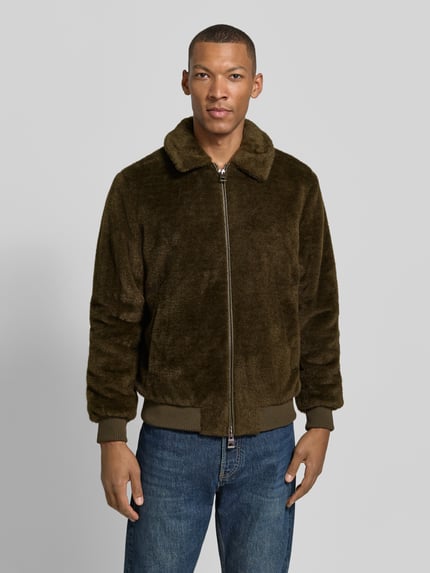 Męski model nosi Regular Fit Bomberjacke mit Zweiwege-Reißverschluss od GUESS, kolor brązowy, tekstura futrzana, kołnierzyk typu kent, wpuszczane kieszenie, prążkowane wykończenia, długość tyłu w rozmiarze M: 66 cm, szerokość ramion w rozmiarze M: 43 cm, szerokość klatki piersiowej w rozmiarze M: 57 cm, długość rękawów w rozmiarze M: 61 cm, widok z przodu całej sylwetki.
