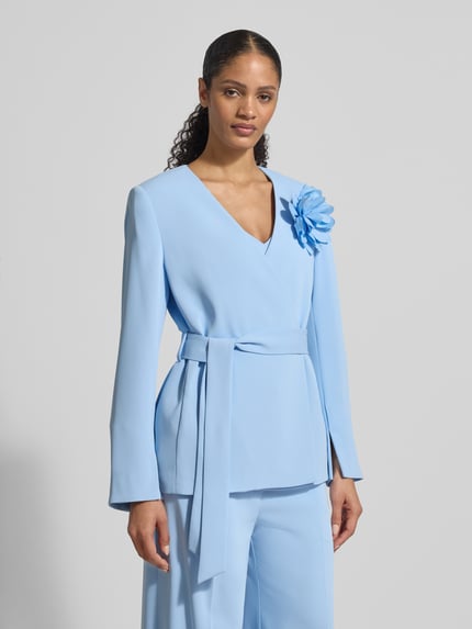 Modelka kobieca nosi Blazer mit Taillengürtel od Christian Berg Cocktail, taliowany krój, kopertowy styl z dekoltem w serek, ozdobna aplikacja kwiatowa, odpinany pasek w talii, długość tyłu w rozmiarze 36: 68 cm, widok całej sylwetki z przodu.