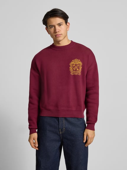 Männermodel trägt bordeauxroten Strickpullover mit Emblem und Rundhalsausschnitt von PROHIBITED, reine Baumwolle, Relaxed Fit, Ganzkörperansicht von vorne.
