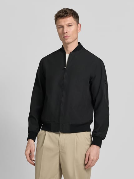Męski model nosi bomberka z mieszanki materiałów z dodatkiem streczu Jack & Jones Premium, model 'BLAASPEN', stójka, zamek błyskawiczny, wpuszczane kieszenie, elastyczne wykończenia, czarna, luźny krój, długość tyłu w rozmiarze M 65 cm, szerokość ramion w rozmiarze M 48 cm, szerokość klatki piersiowej w rozmiarze M 60 cm, długość rękawów w rozmiarze M 66 cm, widok na cały przód.