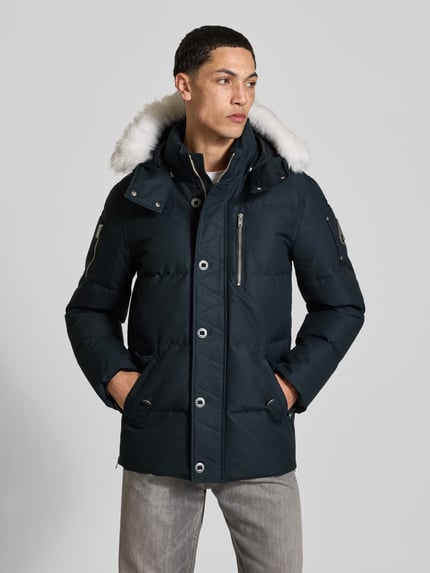 Männermodel trägt Daunenjacke mit abnehmbarer Fellkapuze von Moose Knuckles in Unifarbe, strukturierte Webware mit Steppeinsatz, Ganzkörperansicht von vorne