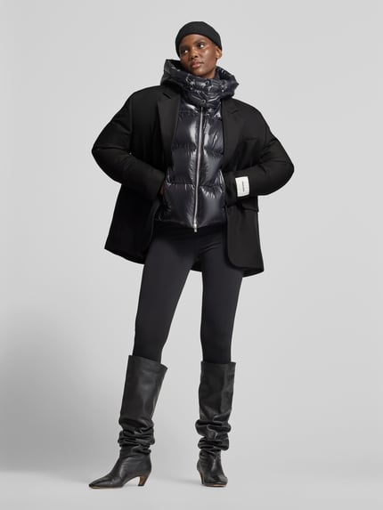 Frauenmodel trägt Regular Fit Longblazer mit Label-Detail von black palms in Schwarz, kombiniert mit Steppjacke, Ganzkörperansicht von vorne. Optik unifarben, Reverskragen, 1-Knopf-Verschluss, Langarm, Pattentaschen.