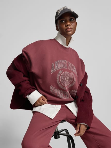 Frauenmodel trägt Sweatshirt mit Logo-Print Modell 'MILES SWEATSHIRT' von Anine Bing in Bordeaux mit unifarbener Optik, Motiv-Print, langem Arm und rundem Kragen, gerippte Bündchen, Oversize-Stil, Ganzkörperansicht von vorne.