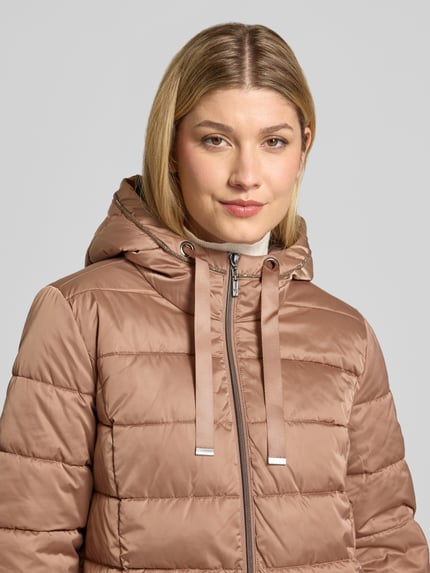 Frauenmodel trägt Steppmantel mit Reißverschlusstaschen von monari in Beige, sichtbar glatte Textur und Kapuze, Nahaufnahme Oberkörper.
