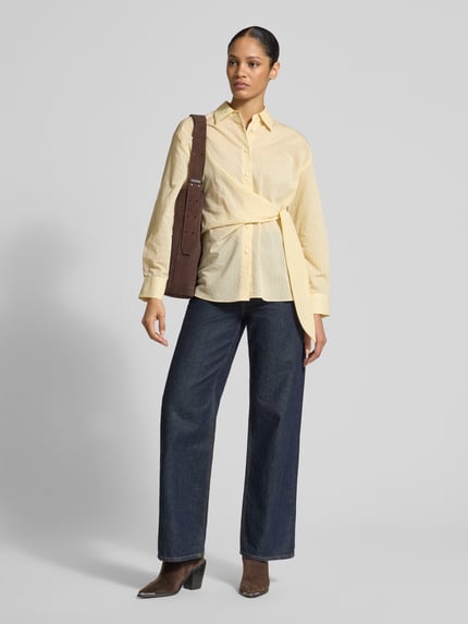 Frauenmodel trägt Regular Fit Bluse mit Knotendetail Modell 'CHIPITI' von Mango aus Baumwoll-Mix, Hemdblusenkragen, Knopfleiste, Rückenlänge 56 cm, Ärmellänge 68 cm, gelb, mit strukturierter Oberfläche, Ganzkörperansicht von vorne.