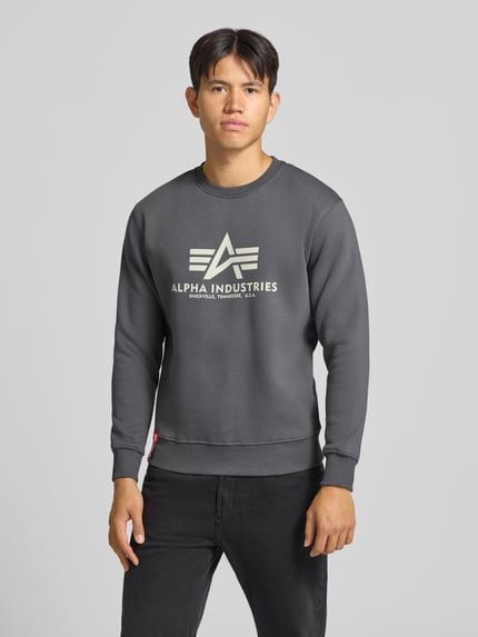 Mężczyzna nosi Regular Fit Sweatshirt aus Baumwoll-Mix mit Logo-Print von Alpha Industries, ciemnoszary, nadruk z logo, mieszanka bawełny, krój regular fit, plisa, długi rękaw, prążkowane wykończenia, okrągły dekolt, długość tyłu w rozmiarze M: 67 cm, długość rękawów w rozmiarze M: 64 cm, zdjęcie w całej sylwetce od przodu.