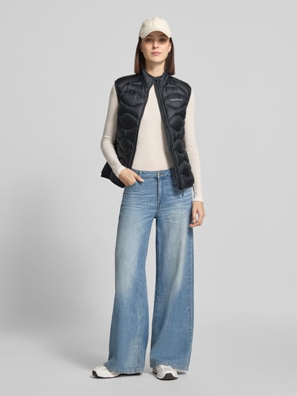 Frauenmodel trägt Steppweste mit Daunen-Federn-Füllung Modell 'HELIUM' von Peak Performance in Schwarz, kombiniert mit weiter Jeans und weißen Sneakers, Stehkragen und Reißverschluss, Slim Fit, Ganzkörperansicht von vorne.