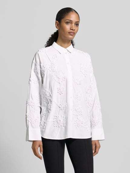 Vrouwenmodel draagt Relaxed Fit Hemdbluse floralem Allover-Besatz van Jake*s Collectie, wit met bloemenmotief, overhemkraag, knoopsluiting, sportmanchetten, ruglengte 70 cm, mouwlengte 50 cm, geheel van voren gefotografeerd.