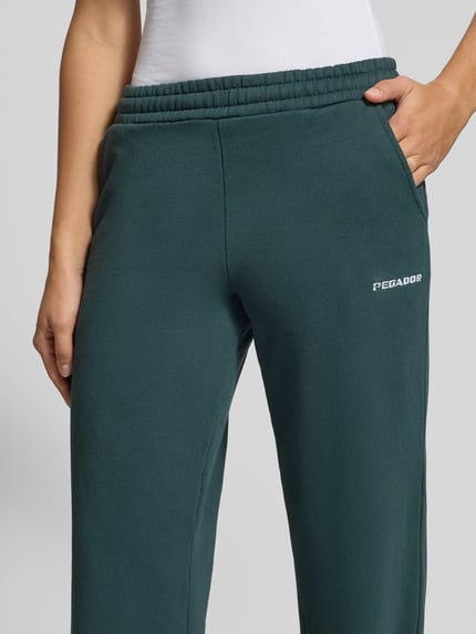 Kobiece model prezentuje Flared Fit Jogginghose aus Baumwoll-Mix Model 'Anni' von PEGADOR, zielony kolor, wpuszczane kieszenie, wyhaftowane logo, elastyczny pas, długość wewnętrzna nogawki w rozmiarze S: 82 cm, mieszanka bawełny, detaliczne ujęcie przodu.
