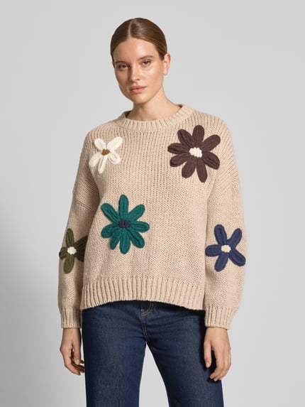 Kobiece modelka nosi Relaxed Fit Strickpullover mit Blumen-Stickerei od Apricot, Imitacja dzianiny, krój relaxed fit, kwiatowy haft, opuszczone ramiona, krój z długim rękawem, prążkowane wykończenia, prążkowana plisa, okrągły dekolt, Długość tyłu w rozmiarze S: 57 cm, Długość rękawów w rozmiarze S: 44 cm, jasny beż z kolorowymi kwiatami, widok całej sylwetki z przodu.