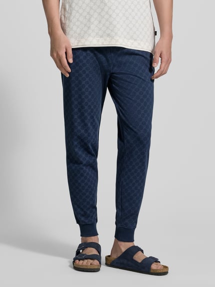 Mężczyzna nosi Sweatpants mit Logo-Muster und elastischem Bund od JOOP! Collection, granatowe, bawełna, krój regular fit, wzór logo, elastyczny pas, długość nogawki wewnętrznej w rozmiarze M: 75 cm, całkowity widok z przodu.