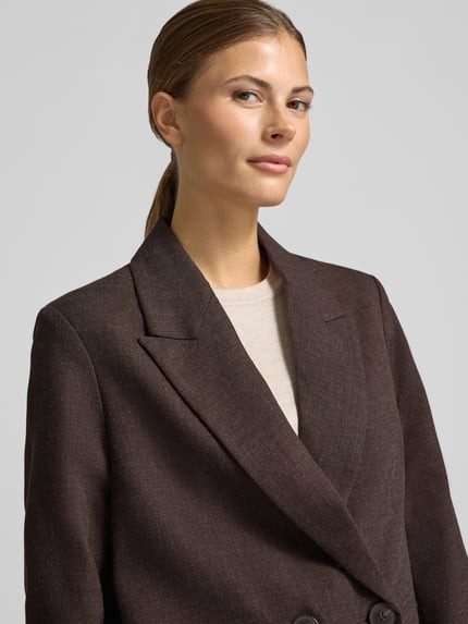 Frauenmodel trägt Loose Fit Longline Blazer mit Schulterpolstern von VERO MODA in Dunkelbraun, Reverskragen, Knöpfe, Webware, Nahaufnahme Oberkörper.