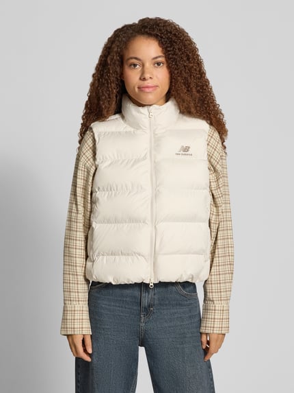 Frauenmodel trägt Steppweste mit Stehkragen Modell 'BOSTON PUFFER VEST' von NEW BALANCE, unifarben, strukturierte Webware, Logo-Print, Reißverschlusstaschen, Ganzkörperansicht von vorne.