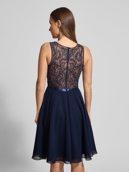 Kobiece modelka nosi sukienkę koktajlową z obszyciem z kamieni stras Cocktailkleid mit Strasssteinbesatz od LUXUAR FASHION, półprzezroczysty materiał, lekko taliowany fason, długość do kolan, jednokolorowy design, zamek błyskawiczny, okrągły dekolt, długość tyłu 96 cm, szerokość klatki piersiowej 41 cm, widok całej sylwetki z tyłu.