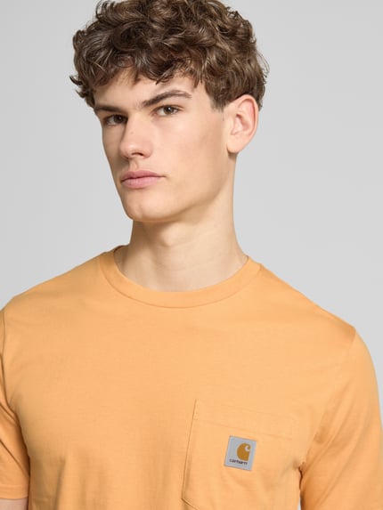 Männermodel trägt Regular Fit T-Shirt mit Logo-Patch von Carhartt Work In Progress in Orange, Rundhalsausschnitt, Nahaufnahme Oberkörper.