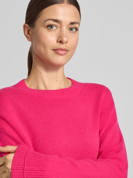 Modelka kobieca nosi Strickpullover mit überschnittenen Schultern od Christian Berg Woman w kolorze intensywnego różu, luźny krój, okrągły dekolt, prążkowane wykończenia, długi rękaw, faktura dzianiny, długość tyłu w rozmiarze M: 60 cm, długość rękawa w rozmiarze M: 58 cm, zbliżenie na górną część ciała.