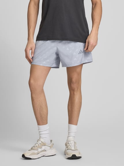 Männermodel trägt Regular Fit Shorts mit Logo-Allover-Muster Modell 'WE BRLO SHO' von adidas Training in Optik: Unifarben, aus Satin, elastischer Bundabschluss und seitlicher Reißverschlusstasche, Ganzkörperansicht von vorne.
