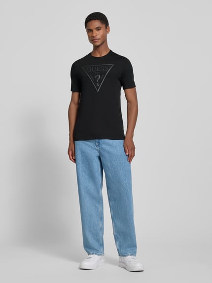Männermodel trägt T-Shirt mit Logo und Rundhalsausschnitt von GUESS in Schwarz, Reine Baumwolle, Slim-Fit, Rückenlänge 70 cm, Ganzkörperansicht von vorne.
