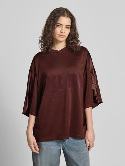 Frauenmodel trägt Oversized T-Shirt mit Label-Print von Karo Kauer in Dunkelbraun, V-Ausschnitt, überschnittene Schultern, Ganzkörperansicht von vorne.