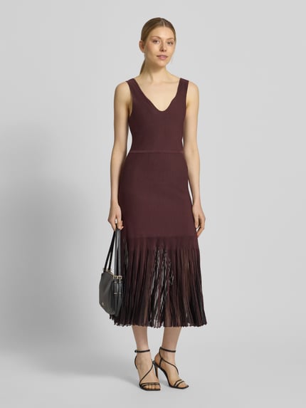 Frauenmodel trägt Strickkleid aus Rippstrick von BOSS in Dunkelrot mit V-Ausschnitt und transparenten Plisseefalten, Ganzkörperansicht von vorne.