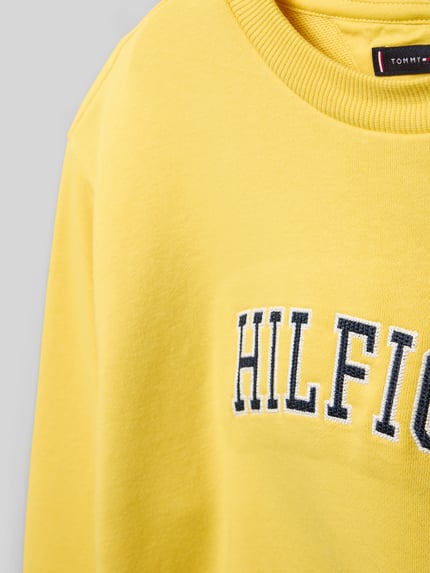 Dziewczynka nosi Regular Fit Sweatshirt aus Baumwoll-Mix od Tommy Hilfiger Teens, materiał bawełniany, kremowy, detal z nadrukiem, crew neck, długi rękaw, wykończenie ściągaczowe, tekstura sweat, detailaufnahme górnej części.