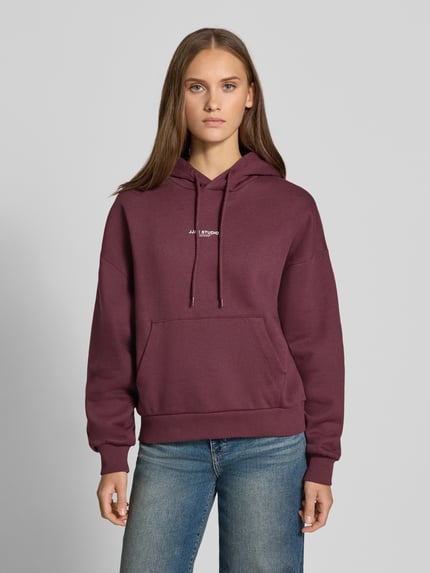 Frauenmodel trägt Relaxed Fit Hoodie aus Baumwoll-Mix Modell 'VESTERBRO' von JJXX in Dunkelrot mit Label-Detail, Känguru-Tasche, Kapuze mit Tunnelzug, Ganzkörperansicht von vorne.