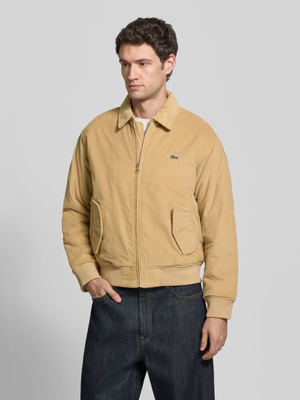 Männermodel trägt Bomberjacke mit Cordstruktur von LACOSTE in Beige, strukturiertes unifarbenes Material, Reißverschluss, seitliche Taschen, Nahaufnahme Oberkörper.