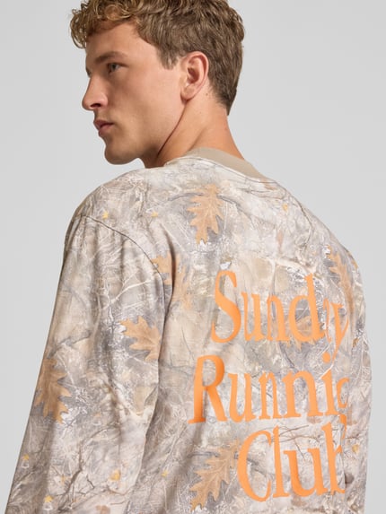 Männermodel trägt Longsleeve mit Logo- und Motiv-Print von ANOTHER COTTON LAB in Naturtönen mit orangefarbenem Schriftzug, Nahaufnahme Rückansicht.