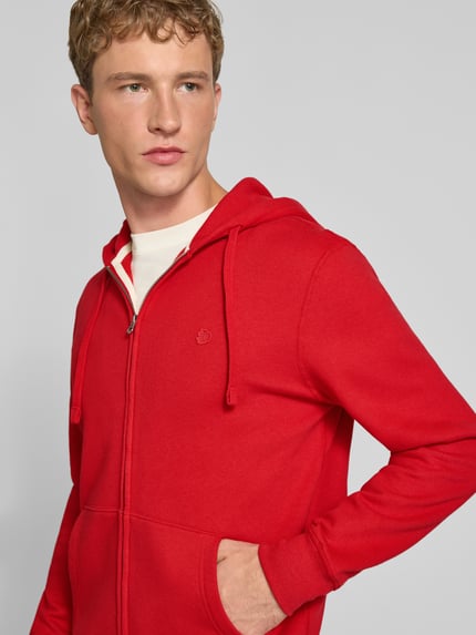 Männermodel trägt Regular Fit Sweatjacke aus Baumwoll-Mix mit Label-Detail von Superdry in Rot, Kängurutasche, Nahaufnahme Oberkörper. Herren Sweatjacke, Kapuze mit Tunnelzug, elastische Abschlüsse, Rückenlänge und Ärmellänge bei Größe M je 65 cm.