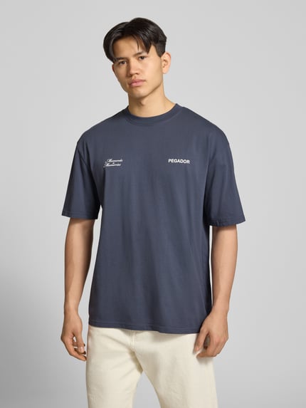 Männermodel trägt Oversized Herren T-Shirt von Pegador aus Reine Baumwolle, dunkelblau, Label-Detail, Ganzkörperansicht von vorne. Modell 'Rochefort', Oversized-Schnitt, Rundhalsausschnitt, Motiv-Print auf der Rückseite, Rückenlänge bei Größe M: 72 cm.