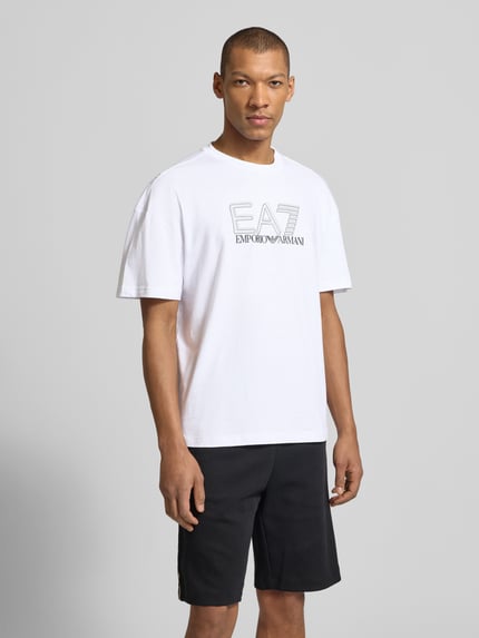 Männermodel trägt T-Shirt mit Logo und Rundhalsausschnitt von EA7 Emporio Armani, reine Baumwolle, Regular-Fit, weiße Farbe, Ganzkörperansicht von vorne.