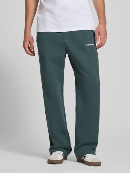 Männermodel trägt Loose Fit Sweatpants aus Baumwoll-Mix mit Label-Detail von PEGADOR, grün, mit Eingrifftaschen, Ganzkörperansicht von vorne.