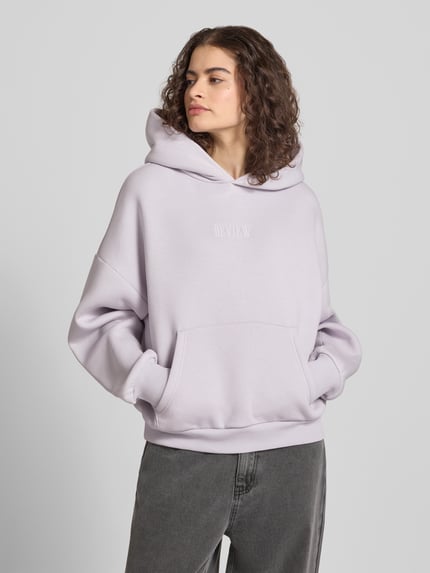 Frauenmodel trägt Relaxed Fit Sweatshirt mit Logo-Print von Review aus Baumwoll-Mix mit Logo-Stitching, Känguru-Tasche, elastische Abschlüsse, Ganzkörperansicht von vorne.