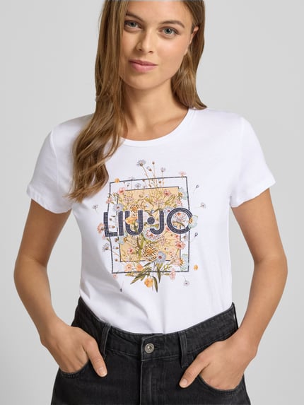 Kobiece modelka nosi T-shirt z nadrukiem z logo i obszyciem ozdobnymi kamieniami od Liu Jo White, bawełna, krój regular fit, okrągły dekolt, długość tyłu 62 cm przy rozmiarze S, widok całego przodu.