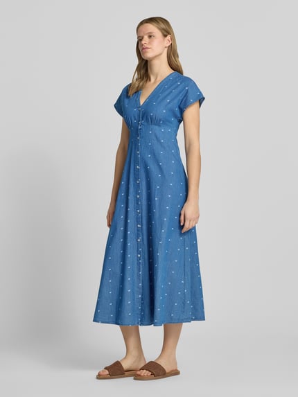 Vrouwenmodel draagt tailliertes jeanskleid uit puur katoen model 'NOVA AZURE' van Only, denimlook, getailleerd, midilengte, V-hals, all-over motief, knoopsluiting, korte mouw, ganzichaamsaanzicht van voren.