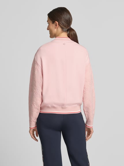 Männermodel trägt Regular Fit Strickjacke aus Mieszanka wiskozy-Mix von Marc Cain, Stójka, zamek błyskawiczny dwustronny, paski w kontrastowym kolorze, długość tyłu w rozmiarze 36: 59 cm, długość rękawów w rozmiarze 36: 58 cm, mieszanka wiskozy i bawełny, kolor jasnoróżowy, tekstura gładka, widoczne detale na rękawach, widok całej sylwetki z tyłu.