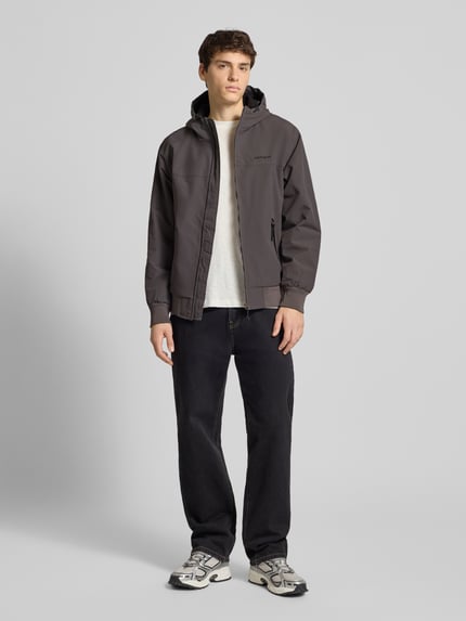 Männermodel trägt Jacke mit Kapuze von Carhartt Work In Progress in Dunkelgrau, Regular Fit, Reißverschluss, Label-Stitching, gerippte Abschlüsse, Reißverschlusstaschen, Ganzkörperansicht von vorne