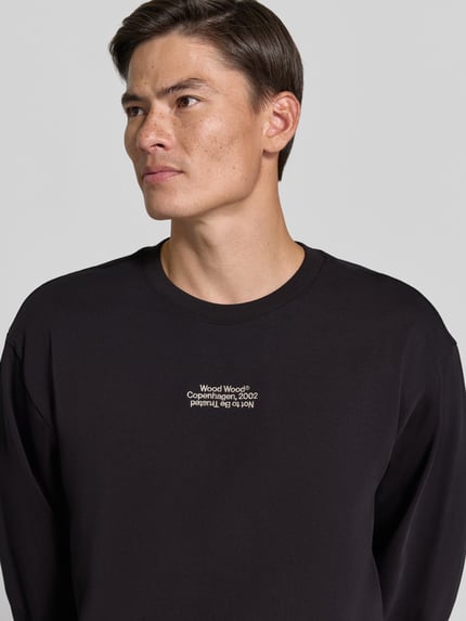 Mężczyznamodel nosi czarną bluzkę z długim rękawem Regular Fit Longsleeve aus reiner Baumwolle Model 'Ryan' od Wood Wood, wyhaftowane logo, bawełna, krój regular fit, długość tyłu w rozmiarze M: 79 cm, ujęcie: zbliżenie górnej części ciała.