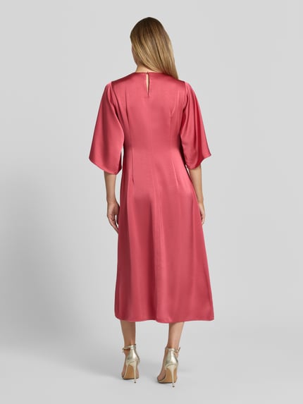 Modelka prezentuje sukienkę midi z satyny o prostym kroju, dekolcie w serek, rękawach-motylkach, model 'Remara' od Neo Noir, długość tyłu 109 cm, szerokość w rozmiarze 36: 47 cm, długość rękawów 38 cm, widok całej sylwetki z tyłu.