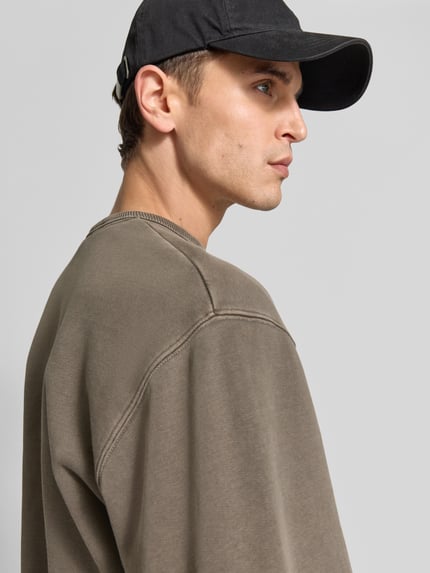 Männermodel trägt Sweatshirt mit Label-Stitching von Forét aus bawełny ekologicznej, prosty krój, okrągły dekolt, prążkowane wykończenia, Długość tyłu w rozmiarze M: 68 cm, zbliżenie górnej części ciała z przodu