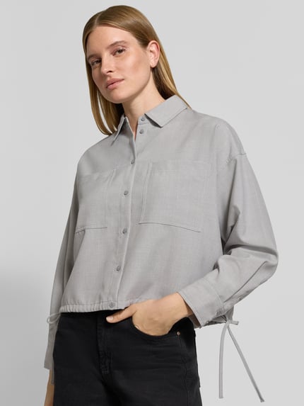 Frauenmodel trägt Cropped Langarmbluse mit Tunnelzugsaum Modell 'Felanga' von OPUS in hellgrauer Melange-Optik mit Strukturmuster, Knopfleiste, Kordeln und Brusttaschen, aus Webware, Nahaufnahme Oberkörper.