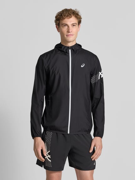 Männermodel trägt schwarze Regular Fit Jacke mit Logo-Print Modell 'ICON' von ASICS TIGER, mit Kapuze, Reißverschluss, elastischen Abschlüssen, Ganzkörperansicht von vorne.